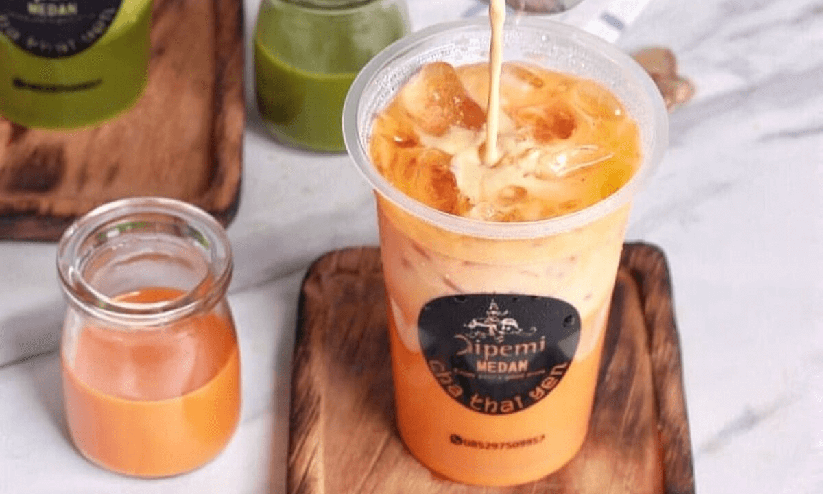 Gelas Thai Tea yang disajikan dengan es batu dan lapisan susu kental manis di atasnya, menampilkan warna oranye yang khas dan menarik, sempurna untuk hari yang panas